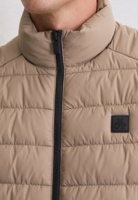 Beige Steppweste mit hohem Kragen, schwarzem Reißverschluss und dezentem Steppmuster. Verfügt über einen kleinen schwarzen Logopatch auf der Vorderseite.