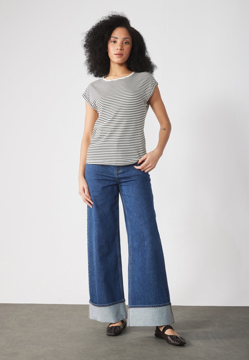 Stripet skjorte med korte ermer i svart og hvitt, kombinert med vide blå denim jeans med dobbeltlagd søm. Svarte flate sko.