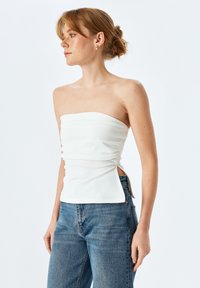 Top blanco sin tirantes con textura fruncida, corsé ajustado y aberturas laterales. Combinado con vaqueros de talle alto azules. Fondo claro.