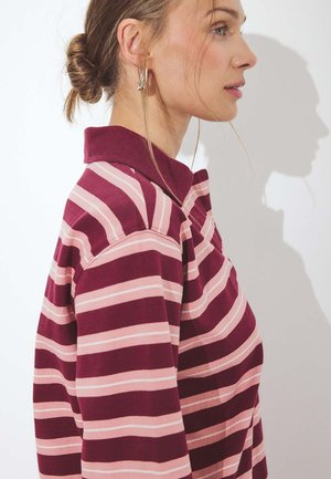 Donna con camicia a righe bordeaux e rosa con colletto, capelli raccolti in uno chignon, rivolta verso destra con un'ombra su sfondo bianco.