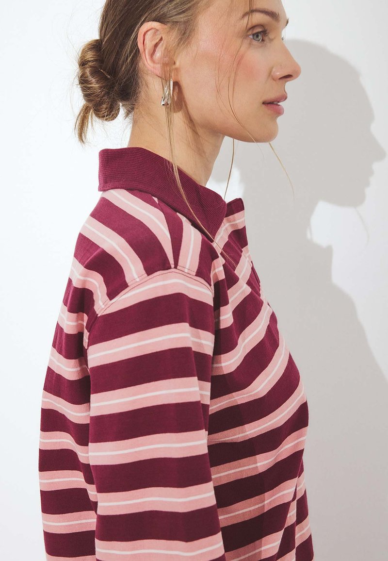 Donna con camicia a righe bordeaux e rosa con colletto, capelli raccolti in uno chignon, rivolta verso destra con un'ombra su sfondo bianco.