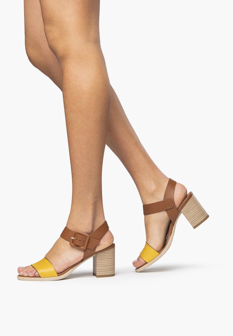 Sandalias de tacón alto abiertas en la punta de color marrón y amarillo, con una correa de hebilla y un tacón de bloque texturizado, que presentan un material similar al cuero suave.