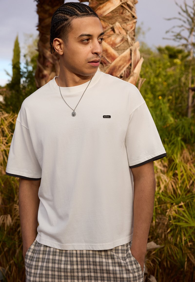 White short-sleeve t-shirt with a black trim and small logo. Paired with plaid shorts in beige, brown, and white hues.
