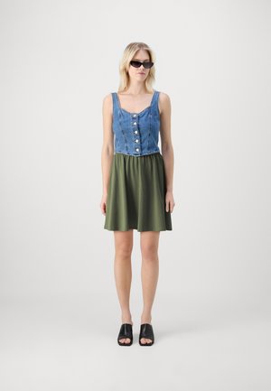 Džínový crop top s detailním zapínáním, kombinovaný se zelenou sukní. Modelka má na sobě černé skládací sandály a sluneční brýle ve stylu kočičích očí. Jednoduché pozadí.