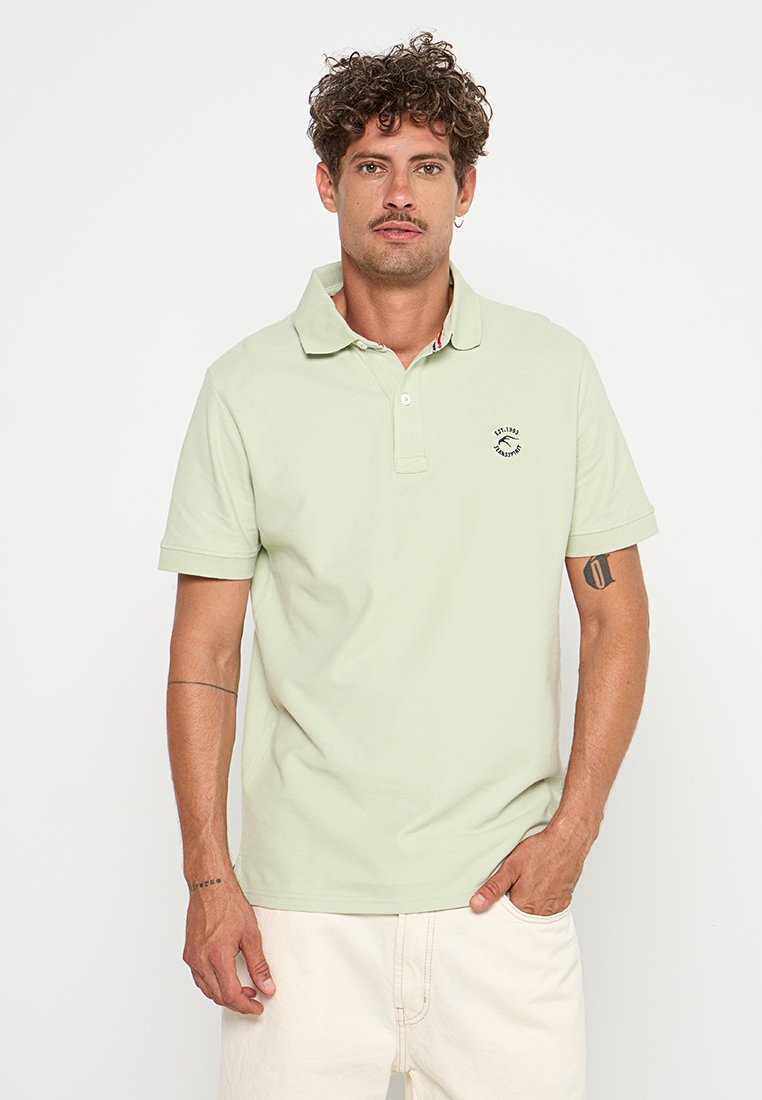 Helles grünes Poloshirt aus Baumwolle mit einem Kragen und kurzen Ärmeln. Auf der linken Brust befindet sich ein kleines Logo. Einfaches Design, glatte Textur.