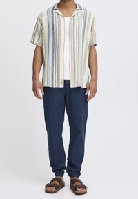 Chemise à manches courtes avec col, rayures verticales jaunes, bleues et grises, associée à un pantalon fuselé bleu marine et des sandales marron à brides réglables.