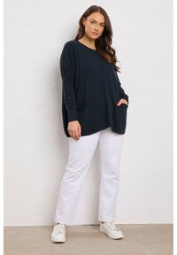 Maglione oversize di colore navy con maniche lunghe, tasche laterali e scollatura rotonda, abbinato a pantaloni bianchi e sneakers bianche.