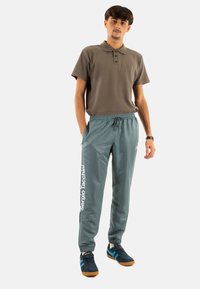 Grijze sportbroek met elastische tailleband en wit "Sergio Tacchini" logo. Gepaard met een bruin poloshirt met korte mouwen en marineblauwe schoenen.