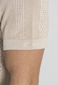 Beige gebreide sweater met textuurpatronen, open weefdesigns en een geribde manchette met het "Gianni Kavanagh" logo geborduurd op de zoom.