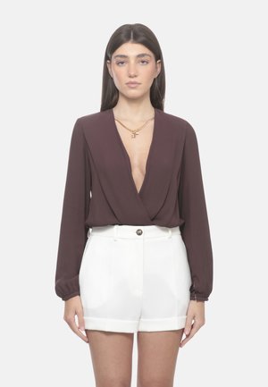 Donna che indossa una camicetta marrone a maniche lunghe con scollo a V profondo, pantaloncini bianchi a vita alta con bottone e una collana a catena dorata.