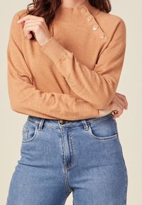 Světlý hnědý svetr s vysokým límcem, zdobený knoflíky na ramenech. Kombinován s klasickými modrými džínami. Měkká textura, úzký střih.