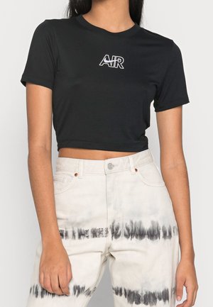Femme portant un t-shirt noir court avec le logo "AIR" et un jean blanc taille haute avec des motifs de tie-dye noir autour des cuisses et des mollets.