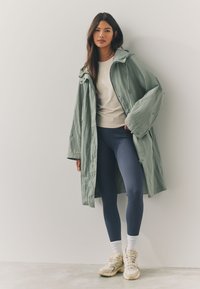 Long, light green trench coat over a white top, paired with navy ribbed leggings and beige sneakers. The coat features a relaxed fit and drawstring.