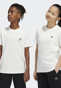 Deux enfants portant des t-shirts blancs unis avec un logo Adidas noir sur la poitrine, associés à un pantalon noir. Design simple à col rond, manches courtes.