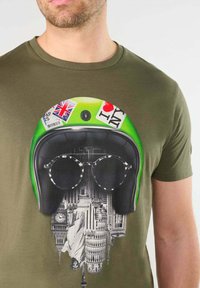 Camiseta verde oliva con un gráfico de un casco vintage adornado con pegatinas y grandes gafas redondas, que muestra paisajes urbanos debajo.