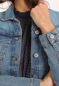 Giacca di jeans azzurro chiaro con bottoni d'argento, caratterizzata da apertura frontale, colletto appuntito e dettagli di cuciture arancioni su una camicetta scura a pois.