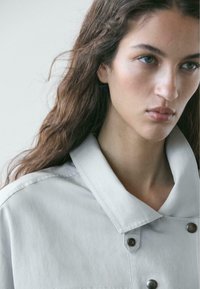 Giacca grigio chiaro con ampio colletto, chiusura con bottoni e texture liscia. Il tessuto sembra morbido e leggero.