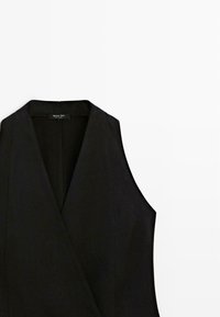 Černá vesta bez rukávů s hlubokým výstřihem do V a přizpůsobeným střihem, rozložená na bílém pozadí.