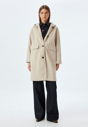 Abrigo de lana beige con capucha, dos bolsillos frontales y botones marrones, combinado con pantalones de mezclilla oscuros de pierna ancha y zapatos marrones de punta.