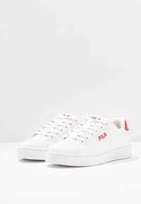 Sneakers in pelle bianca con dettagli rossi e superficie testurizzata, dotate di lacci frontali e suola di gomma spessa. Logo Fila in evidenza.