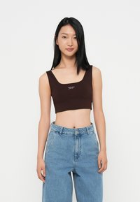 Brun cropped topp med firkantet halsutringning og brede stropper, med en logo i midten. Kombinert med lyseblå høytlivsjeans.