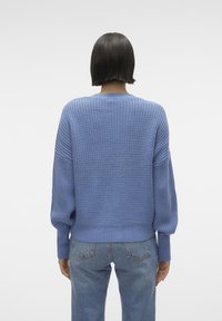Jasnoniebieski sweter dziergany z teksturowanym wzorem, opuszczonymi ramionami i ściągaczami na mankietach. Noszony z niebieskimi dżinsami, prezentujący luźny krój.