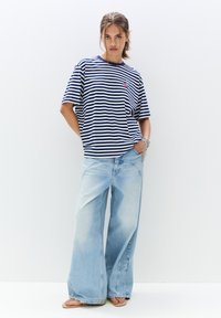 Marineblå og hvide striber oversized T-shirt parret med lyseblå brede jeans. Modellen står med hænderne i lommerne og har flip-flops på.