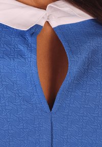 Top texturizado azul con cuello blanco y una apertura en forma de lágrima en la parte posterior, con un cierre de botón visible.