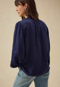 Blusa a maniche lunghe blu navy con colletto elasticizzato, spalle arricciate e dettagli ricamati sulle maniche, abbinata a jeans in denim chiaro.