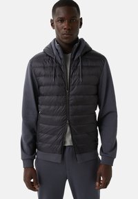 Mann trägt eine schwarze gesteppte ärmellose Jacke über einem grauen Hoodie und passenden grauen Jogginghosen, steht vor einem schlichten Hintergrund.