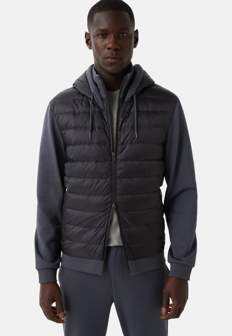 Mann trägt eine schwarze gesteppte ärmellose Jacke über einem grauen Hoodie und passenden grauen Jogginghosen, steht vor einem schlichten Hintergrund.