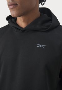 Svart ärmlös hoodie med en slät yta, försedd med en liten grå logotyp på bröstet. Tyget verkar lätt och elastiskt.