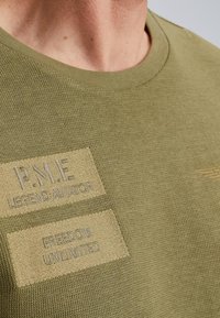 Tee s kratkim rukavima od maslinaste pamuka s teksturiranim pletenjem. Ima dva ušivena flastera: jedan bež s natpisom "P.M.E LEGEND AVIATOR" i drugi s natpisom "FREEDOM UNLIMITED."