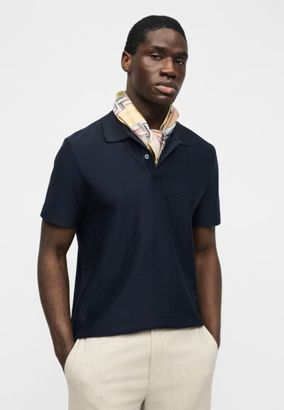 Άνδρας που φοράει μπλε ναυτικό polo με κοντά μανίκια, μπεζ παντελόνι και πολύχρωμο φουλάρι με σχέδιο γύρω από το λαιμό, όρθιος με τα χέρια στις τσέπες.