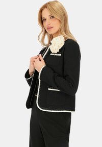 Czarny żakiet z białymi wykończeniami, teksturowany materiał, dwie przednie kieszenie oraz dekoracyjny kremowy kwiatowy akcent na ramieniu. Dopasowany krój.