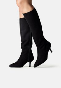 Cizme negre din suede, până la genunchi, cu vârfuri ascuțite și tocuri subțiri de înălțime medie, purtate pe picioare într-un fundal simplu, deschis la culoare.