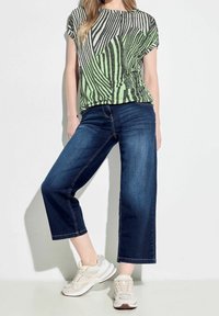 Grün und schwarz gestreiftes T-Shirt mit kurzen Ärmeln, kombiniert mit dunkelblauen weit geschnittenen Jeans und hellen Sneakers. Glattes Material und lockere Passform.