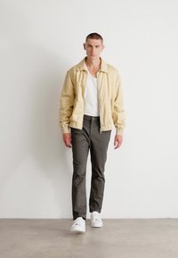 Beige lichtgewicht bomberjack, ribbelkraag, witte ribbel-tanktop, donkere broek en witte sneakers. Model staat tegen een eenvoudige achtergrond.