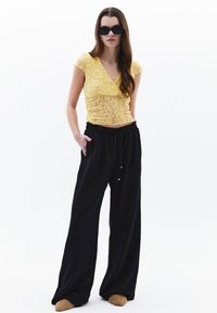 Top corto de encaje amarillo con escote en V, combinado con pantalones negros holgados y zapatos deslizantes con estampado. Accesorios y peinado sencillos.