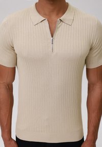 Beige Polo-Shirt mit kurzen Ärmeln und geripptem Textur, mit einem Reißverschluss vorne und einem klassischen Kragen. Keine sichtbaren Muster oder Verzierungen.