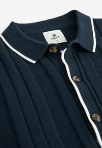 Laivastonsininen polo-paita, jossa on teksturoitu kohta, valkoinen kaulusreunus ja kaksi ruskeaa nappia nappilistalla. Paidassa on myös etiketti, jossa lukee "NEXT" ja koko M.