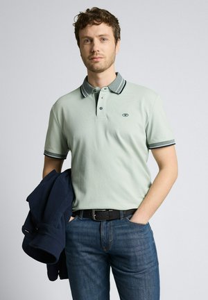 Jonge man met kort krullend haar draagt een lichtgroen poloshirt en blauwe spijkerbroek, houdt een marineblauwe trui vast en staat tegen een effen achtergrond.