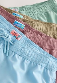 Quatre paires de shorts de bain empilées. Les couleurs incluent le bleu clair, le vert, le marron et le rose. Chaque short possède une ceinture élastique avec un cordon de serrage.