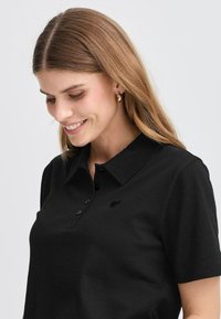 Juodas polo marškinėliai su klasikiniu apykakle, turintys trijų mygtukų užsegimą ir subtilų logotipą kairėje pusėje. Minkšta tekstūra ir prigludęs dizainas.