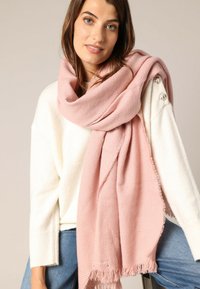 Écharpe tissée rose avec un motif en chevrons, aux bords effilochés et à la texture douce, drapée de manière décontractée. Portée sur un pull blanc.