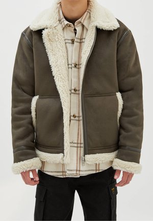 Homme portant une veste en peau retournée marron avec une doublure en laine crème sur une chemise à carreaux beige boutonnée et un pantalon cargo noir.