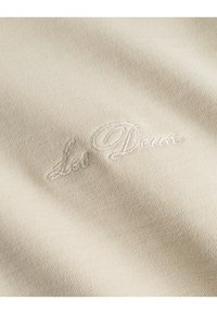 Beige színű szőtt anyag, amely finom textúrával rendelkezik, és a "Les Deux" hímzett logó világos árnyalatban, középre helyezve található.