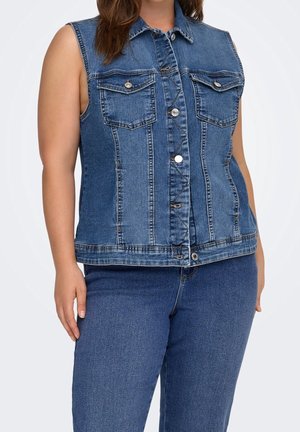 Femme portant un gilet en jean bleu sans manches avec boutons sur le devant et jean bleu assorti sur un fond uni.
