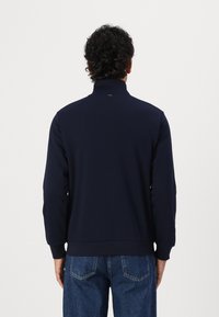 Tummansininen vetoketjullinen huppari, jossa on korkea kaulus ja resorit, valmistettu pehmeästä kankaasta, ominaisuutena yksinkertainen muotoilu ilman näkyviä logoja.