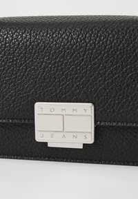 Juodas tekstūruotas odinis krepšys su sidabriniu stačiakampiu užsegimu, išgraviruotu "TOMMY JEANS". Pasižymi elegantišku dizainu ir lygiu paviršiumi.